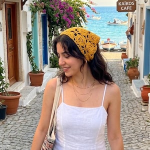 May include: A mustard yellow crochet headscarf with a floral pattern, a white sleeveless midi dress with thin straps, a patterned fabric tote bag, a layered gold chain necklace, a stack of brown beaded bracelets, and a straw hat held in one hand.