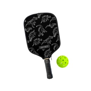 May include: Black pickleball paddle with a white dinosaur pattern and the name "BRITNEY" printed on the paddle. A neon green pickleball is next to the paddle. The paddle handle is black.