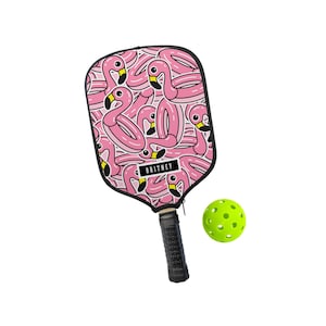 May include: A pink pickleball paddle with a flamingo pattern and the name "BRITNEY" on the paddle face. A green pickleball is next to the paddle. The paddle has a black grip.