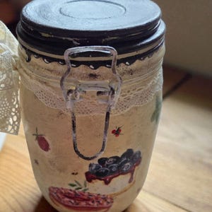 Puede incluir: Un tarro decorativo con tapa negra y cierre metálico. El tarro está pintado con un fondo color crema y presenta ilustraciones de bayas y una tarta. Un ribete de encaje blanco rodea el tarro.