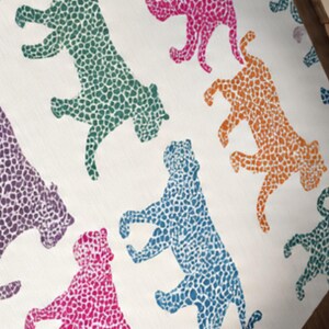 May include: A repeating pattern of colorful leopard silhouettes on a cream-colored background. The leopards are in shades of green, pink, orange, blue, and purple. The design is likely for fabric or wallpaper.