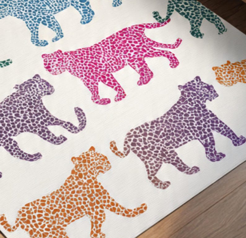 May include: A white rug featuring a repeating pattern of stylized leopard silhouettes in various colors. The leopards are in shades of pink, purple, orange, teal, and green, with a spotted design. The rug is placed on a wooden floor.