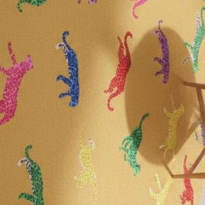 May include: Mustard-colored fabric with a repeating pattern of colorful cheetah silhouettes. The cheetahs are in shades of pink, blue, red, green, yellow, and purple. The fabric has a textured appearance.