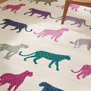 May include: A cream-colored rug featuring a repeating pattern of stylized cheetahs in various colors, including pink, purple, teal, green, and navy. The cheetahs are depicted in a walking pose, with a spotted design. The rug is a decorative home accent.