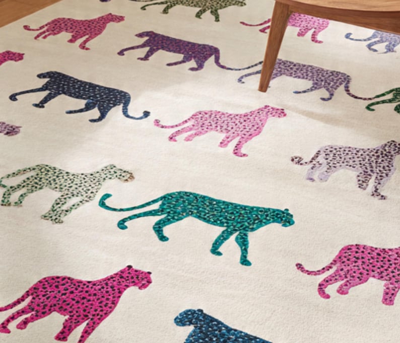 May include: A cream-colored rug featuring a repeating pattern of stylized cheetahs in various colors, including pink, purple, teal, green, and navy. The cheetahs are depicted in a walking pose, with a spotted design. The rug is a decorative home accent.