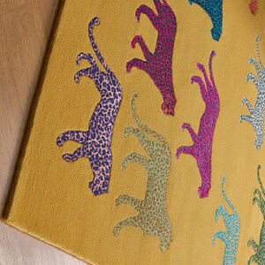 Puede incluir: Alfombra de color amarillo mostaza con un patr&oacute;n repetido de figuras de leopardo estilizadas en azul, rosa y turquesa. Las figuras de leopardo tienen un dise&ntilde;o moteado.
