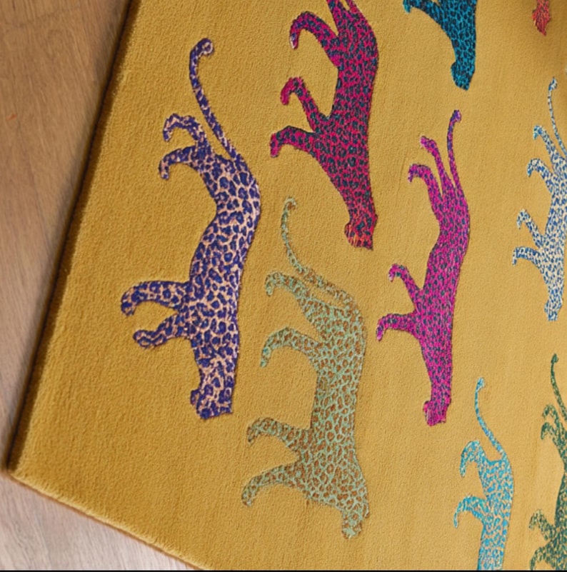 Puede incluir: Alfombra de color amarillo mostaza con un patr&oacute;n repetido de figuras de leopardo estilizadas en azul, rosa y turquesa. Las figuras de leopardo tienen un dise&ntilde;o moteado.