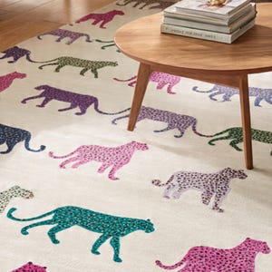 May include: A cream-colored area rug featuring a repeating pattern of stylized cheetahs in various colors, including pink, purple, green, and blue. A round wooden coffee table with books sits on the rug.