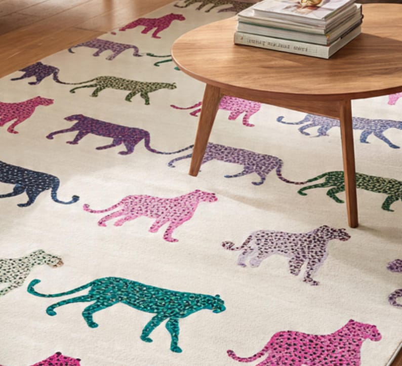May include: A cream-colored area rug featuring a repeating pattern of stylized cheetahs in various colors, including pink, purple, green, and blue. A round wooden coffee table with books sits on the rug.