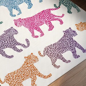 May include: A white rug featuring a repeating pattern of stylized leopard silhouettes in various colors. The leopards are in shades of pink, purple, orange, teal, and green, with a spotted design. The rug is placed on a wooden floor.