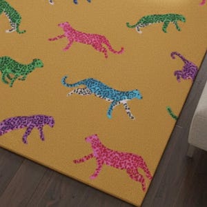 May include: Mustard-colored rug featuring a repeating pattern of colorful cheetahs. The cheetahs are in shades of pink, purple, blue, and green, with a speckled design. The rug is rectangular and appears to be made of a soft material.