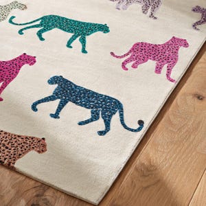 May include: A rectangular area rug with a cream-colored background features a repeating pattern of colorful leopard silhouettes. The leopards are in shades of teal, pink, blue, and brown, with black spots. The rug is placed on a wooden floor.