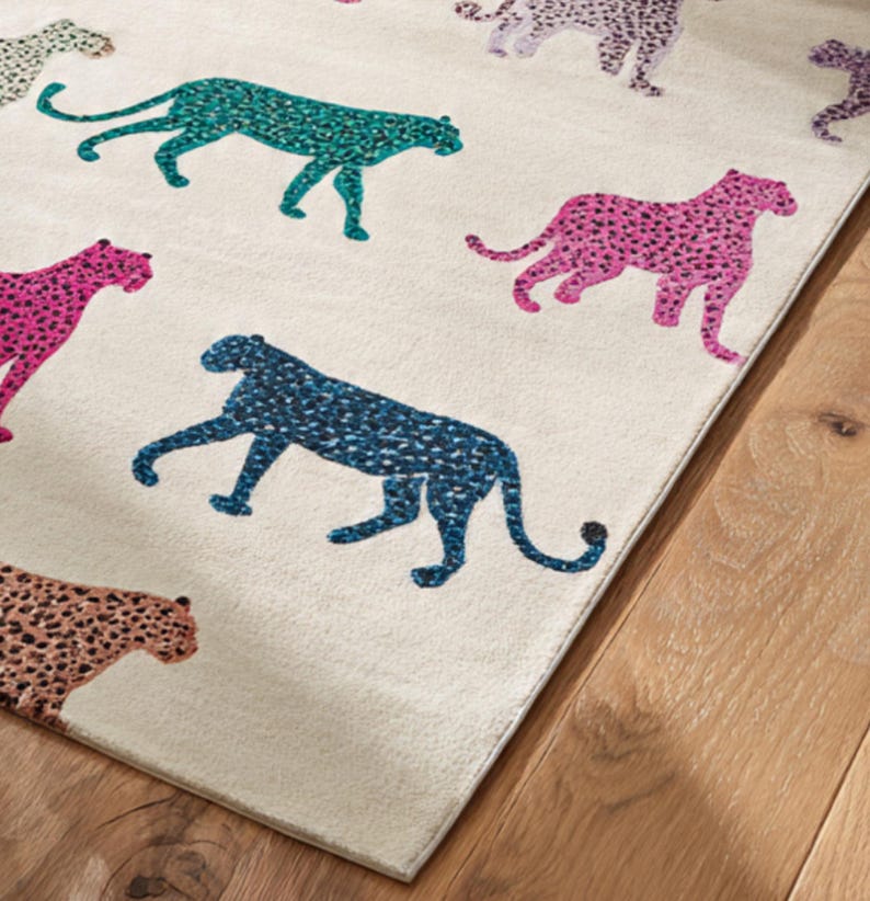 May include: A rectangular area rug with a cream-colored background features a repeating pattern of colorful leopard silhouettes. The leopards are in shades of teal, pink, blue, and brown, with black spots. The rug is placed on a wooden floor.