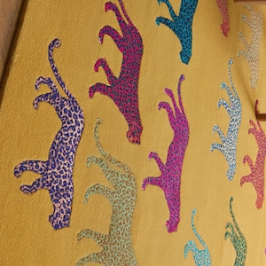 Puede incluir: Tela de color mostaza con un patr&oacute;n repetido de figuras de leopardo estilizadas en varios colores. Los leopardos son de colores vibrantes, incluyendo azul, rosa y verde, con un dise&ntilde;o moteado. Un textil din&aacute;mico y original.