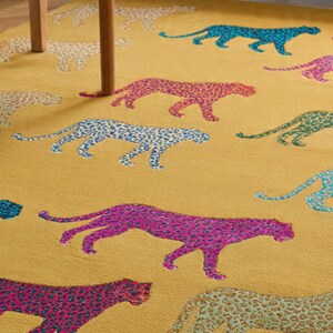 Puede incluir: Alfombra de color amarillo mostaza con un patr&oacute;n repetido de guepardos coloridos. Los guepardos son en tonos de azul, rosa, verde y coral, con un dise&ntilde;o moteado. Se ve una mesa de madera.