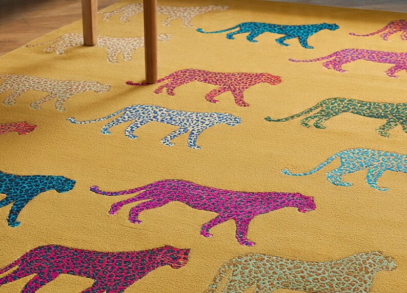 Puede incluir: Alfombra de color amarillo mostaza con un patr&oacute;n repetido de guepardos coloridos. Los guepardos son en tonos de azul, rosa, verde y coral, con un dise&ntilde;o moteado. Se ve una mesa de madera.