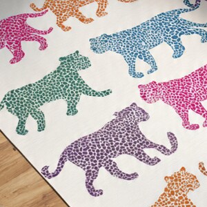 May include: A white textile featuring a repeating pattern of stylized leopard silhouettes in various colors. The leopards are depicted in shades of pink, orange, blue, green, and purple, with a speckled design. The overall aesthetic is playful and vibrant.