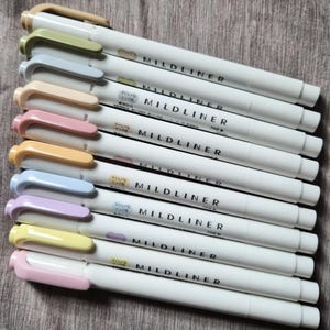 Puede incluir: Una colección de resaltadores Mildliner blancos con tapas de colores pastel. Las tapas son en tonos beige, verde, azul, melocotón, rosa, naranja, azul claro, morado, amarillo y rosa. El nombre de la marca "MILDLINER" está impreso en el lateral de cada bolígrafo.