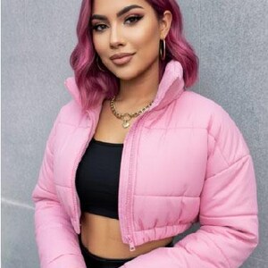 May include: A pale pink, cropped puffer jacket with a high collar and a zip fastening. The jacket is quilted and has long sleeves. The model is wearing a black top and black trousers. Sunglasses are on top of the head.