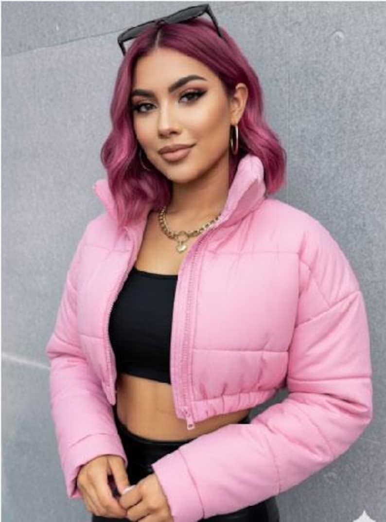 May include: A pale pink, cropped puffer jacket with a high collar and a zip fastening. The jacket is quilted and has long sleeves. The model is wearing a black top and black trousers. Sunglasses are on top of the head.