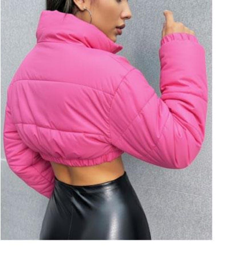 May include: A vibrant pink, cropped puffer jacket with a high collar and long sleeves. The jacket features a quilted design and a fitted shape. The model is wearing a black skirt. The jacket is a striking outerwear item.