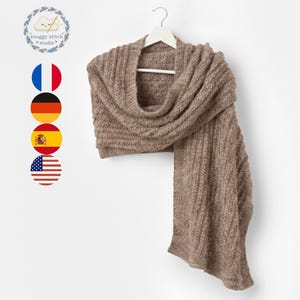 Puede incluir: Bufanda de punto color topo colgada de una percha blanca. La bufanda tiene un diseño texturizado y acanalado y está drapeada para mostrar su longitud y forma. La bufanda está hecha de un material suave y cálido.