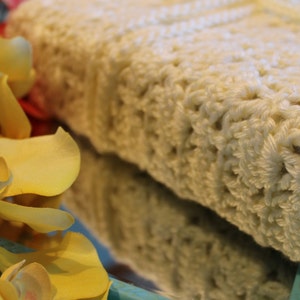 May include: A white crocheted blanket with a delicate pattern. The blanket is folded and displayed on a blue surface with yellow flowers.