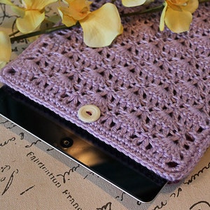 May include: A lavender crocheted tablet case with a white button closure. The case is lying on a surface with a brown background and black writing.