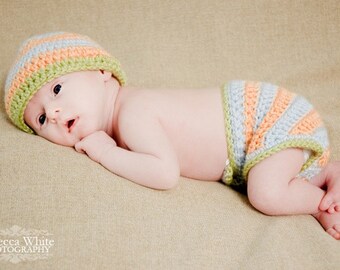 May include: A baby wearing a crocheted hat and nappy cover set. The hat and nappy cover are striped in orange, light blue, and green. The baby is lying on a neutral-coloured surface.