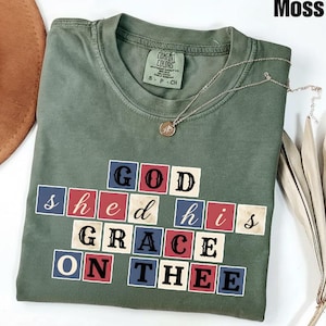 May include: A moss green t-shirt with the phrase "GOD shed his GRACE ON THEE" in a block letter design. A silver necklace with a circular pendant is draped on the shirt. The shirt is folded neatly.