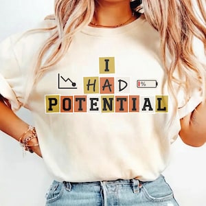 May include: Ivory-colored t-shirt with the text "I HAD POTENTIAL" in a block letter design. The graphic includes a downward trend line and a low battery icon, creating a humorous statement.
