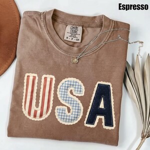 May include: Espresso-coloured t-shirt with the word "USA" appliqu&eacute;d on the front. The "U" has red and white stripes, the "S" is blue gingham, and the "A" is navy blue. A silver necklace with a pendant rests on the shirt.