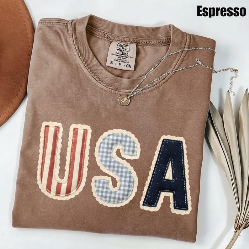 May include: Espresso-coloured t-shirt with the word "USA" appliqu&eacute;d on the front. The "U" has red and white stripes, the "S" is blue gingham, and the "A" is navy blue. A silver necklace with a pendant rests on the shirt.