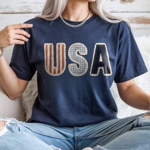 May include: Navy blue t-shirt featuring the word "USA" in a patchwork design. The letters are in red and white stripes, blue gingham, and navy with a white border. The shirt is a relaxed fit and is worn with blue jeans.