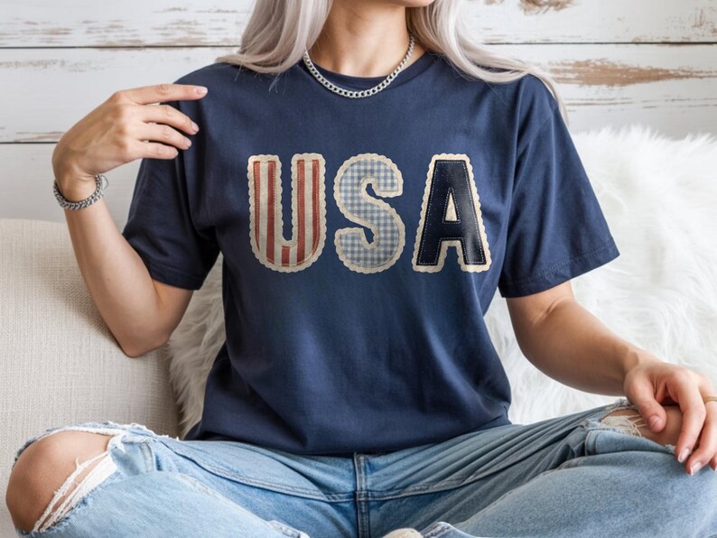 May include: Navy blue t-shirt featuring the word "USA" in a patchwork design. The letters are in red and white stripes, blue gingham, and navy with a white border. The shirt is a relaxed fit and is worn with blue jeans.