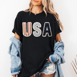 May include: Black t-shirt featuring the word "USA" in a patchwork design. The letters are in red and white stripes, blue gingham, and navy blue. The shirt is worn with a denim jacket and shorts.