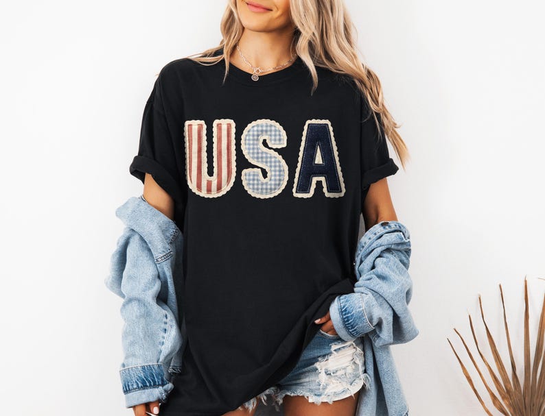 May include: Black t-shirt featuring the word "USA" in a patchwork design. The letters are in red and white stripes, blue gingham, and navy blue. The shirt is worn with a denim jacket and shorts.