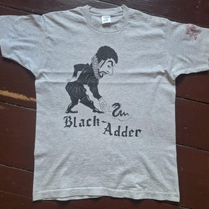 May include: A heather gray t-shirt featuring a black and white illustration of a historical figure with the text "Black Adder". The figure is depicted in a period costume, interacting with a snake.