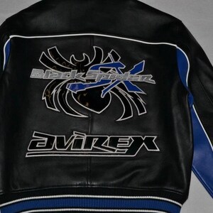 Puede incluir: Chaqueta de cuero negra con detalles blancos y azules. La parte trasera presenta un gran gr&aacute;fico con el texto "Black Spider X" y la marca "Avirex" en letras blancas. La chaqueta tiene rayas azules en las mangas y un dobladillo a rayas azul y blanco.