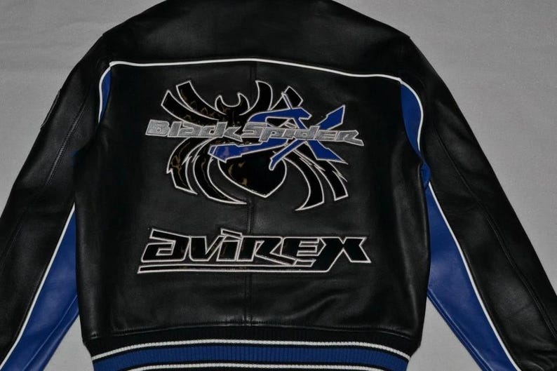 Puede incluir: Chaqueta de cuero negra con detalles blancos y azules. La parte trasera presenta un gran gr&aacute;fico con el texto "Black Spider X" y la marca "Avirex" en letras blancas. La chaqueta tiene rayas azules en las mangas y un dobladillo a rayas azul y blanco.
