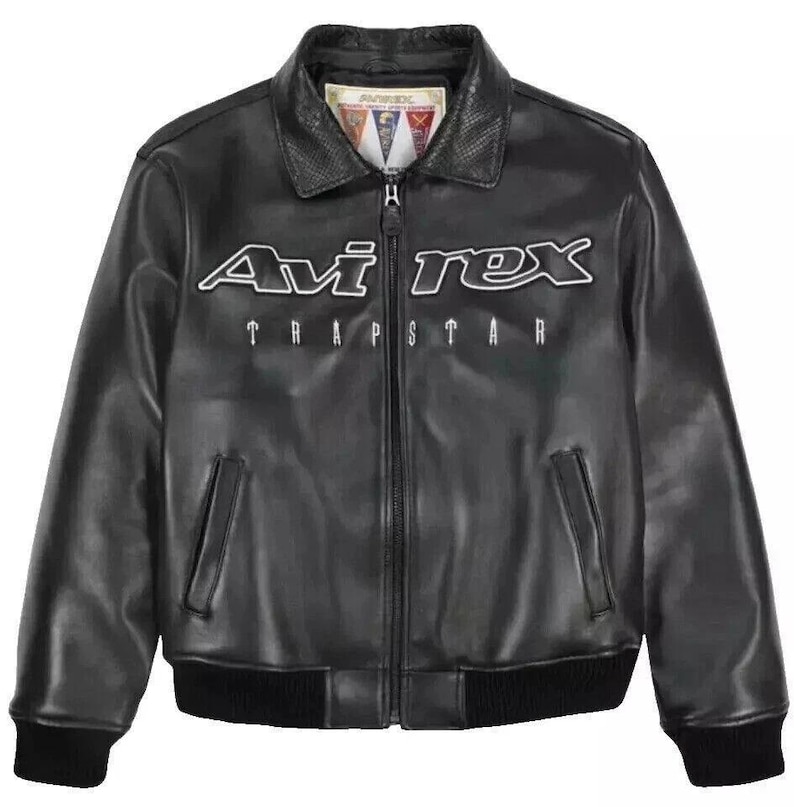 May include: Black leather jacket with the white text "Avi Rex" and "Trapstar" across the chest. The jacket has a collared neckline, a zipper closure, and two front pockets. The cuffs and hem are ribbed.