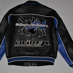 Puede incluir: Chaqueta de cuero negra con detalles azules y blancos. Presenta el texto "Black Spider" y "Avirex" en la espalda. La chaqueta tiene un dobladillo y pu&ntilde;os acanalados con rayas azules y blancas. Medidas en cm.