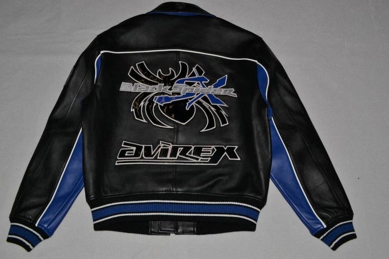 Puede incluir: Chaqueta de cuero negra con detalles azules y blancos. Presenta el texto "Black Spider" y "Avirex" en la espalda. La chaqueta tiene un dobladillo y pu&ntilde;os acanalados con rayas azules y blancas. Medidas en cm.