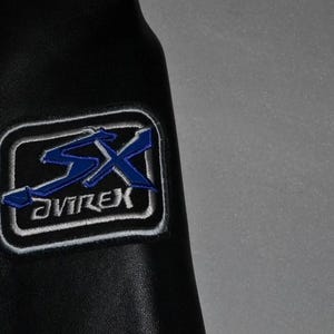 Puede incluir: Detalle de una chaqueta de cuero negra con un parche bordado azul y blanco. El parche presenta las letras "SX" en azul, con la palabra "AVIREX" debajo, todo dentro de un rect&aacute;ngulo con borde blanco.