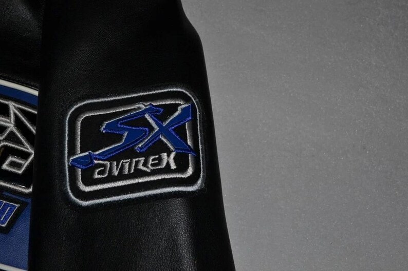 Puede incluir: Detalle de una chaqueta de cuero negra con un parche bordado azul y blanco. El parche presenta las letras "SX" en azul, con la palabra "AVIREX" debajo, todo dentro de un rect&aacute;ngulo con borde blanco.