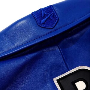 May include: Close-up of a royal blue jacket. The jacket has a small, embroidered emblem on the shoulder and a large letter patch. The material appears to be leather or a leather-like fabric. Stitching is visible along the seams.