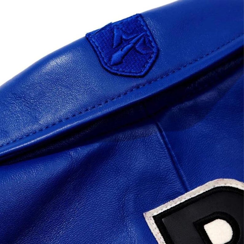 May include: Close-up of a royal blue jacket. The jacket has a small, embroidered emblem on the shoulder and a large letter patch. The material appears to be leather or a leather-like fabric. Stitching is visible along the seams.
