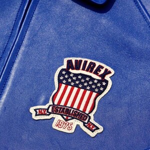 May include: Close-up of a blue leather jacket with a stitched patch. The patch features the word "AVIREX" above a shield design with red and white stripes and stars. The patch also includes the text "ESTABLISHED 1975 NY".