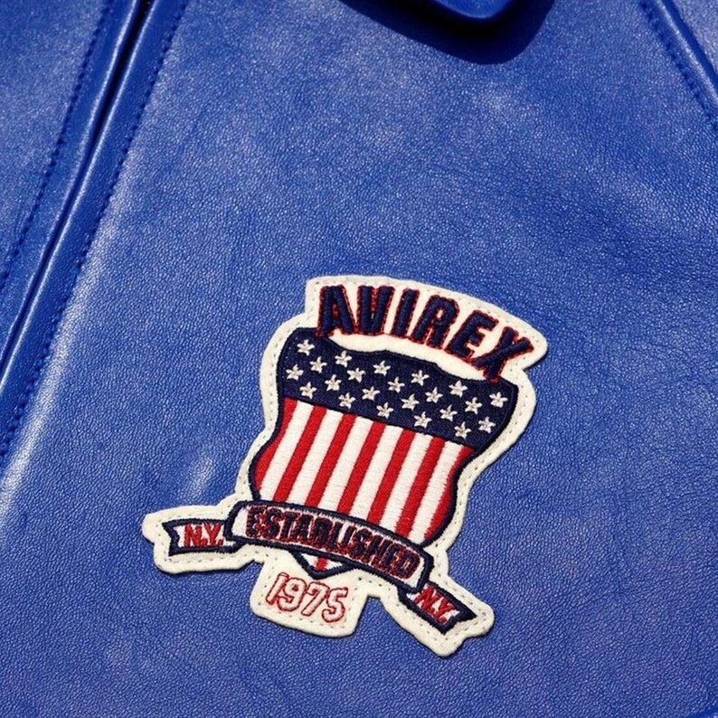May include: Close-up of a blue leather jacket with a stitched patch. The patch features the word "AVIREX" above a shield design with red and white stripes and stars. The patch also includes the text "ESTABLISHED 1975 NY".