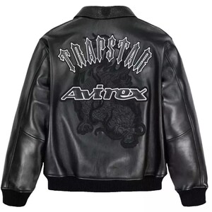 May include: Black leather jacket with a bomber-style design. The back features the text "TRAPSTAR" above a dragon graphic and the "Avirex" logo in white. The jacket has a collar, ribbed cuffs, and a ribbed hem.
